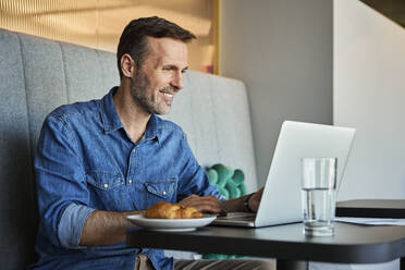 Happy businessman working on laptop in office cafeteria - BSZF02494