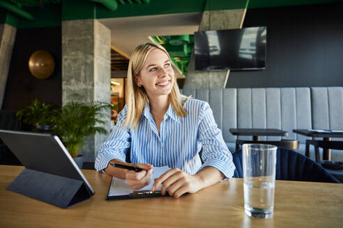 Happy businesswoman sitting with clipboard and tablet PC at desk - BSZF02514