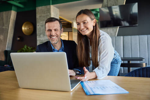 Happy businessman and businesswoman discussing over laptop in office - BSZF02519