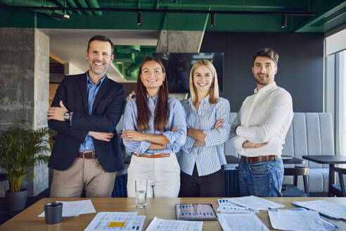 Happy business people standing with arms crossed near graphs at desk - BSZF02533