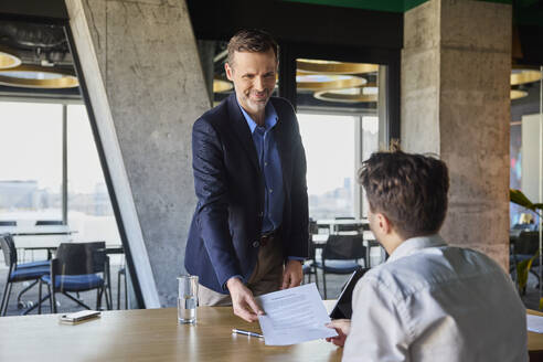 Lächelnder Personalverantwortlicher, der einem Bewerber am Schreibtisch einen Arbeitsvertrag übergibt - BSZF02545