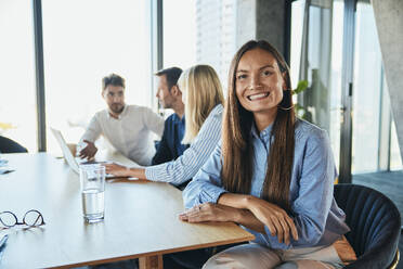 Happy businesswoman sitting with colleagues in background - BSZF02572