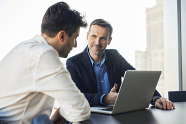 Smiling mature businessman discussing with colleague over laptop at desk - BSZF02582