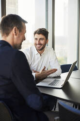 Smiling young businessman discussing with colleague over laptop at desk - BSZF02585