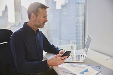 Businessman holding smart phone near laptop and documents at desk - BSZF02590