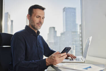 Mature businessman using smart phone near laptop at desk - BSZF02591