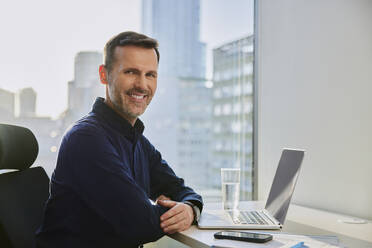 Smiling mature businessman sitting with laptop and smart phone at desk - BSZF02595