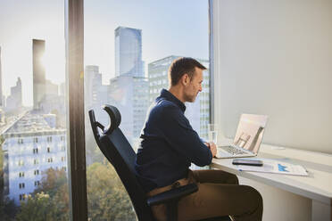 Mature businessman using laptop at desk near window in office - BSZF02598