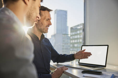 Smiling mature businessman explaining to colleague over laptop at desk in office - BSZF02602