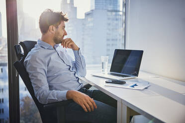 Thoughtful young businessman sitting with laptop at desk - BSZF02605