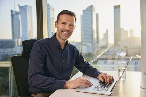 Happy mature businessman with laptop at desk - BSZF02617