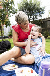 Woman sitting with daughter eating fruits sitting on blanket in garden - WPEF08150