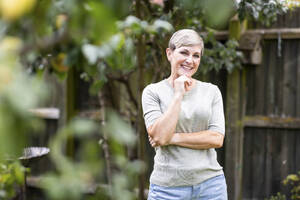 Happy mature woman standing with hand on chin in back yard - WPEF08157