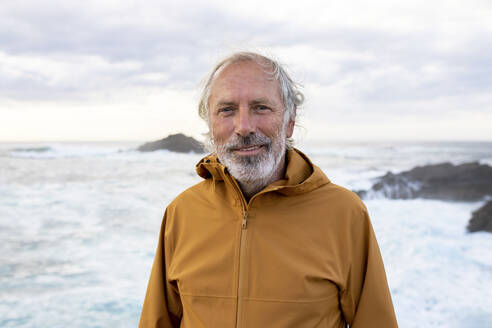 Smiling senior man wearing jacket and standing near sea - MMPF01135