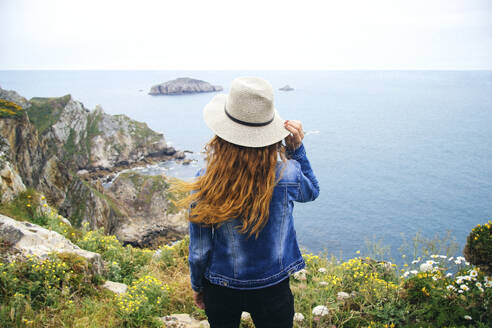 Rothaarige Frau steht auf einer Klippe und schaut auf das Meer in Asturien, Spanien - MMPF01143