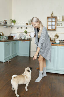 Smiling woman giving snack to Pug dog in kitchen at home - EHAF00192