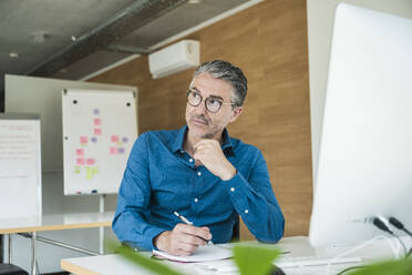 Contemplative businessman holding pen and sitting at desk in office - UUF31046