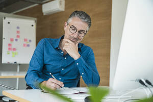 Geschäftsmann mit Hand am Kinn, der am Schreibtisch in sein Tagebuch schreibt - UUF31049