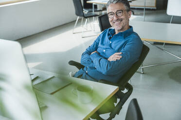 Happy businessman with arms crossed sitting at desk in office - UUF31052