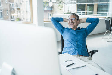 Happy businessman sitting at desk with hands behind head in office - UUF31054