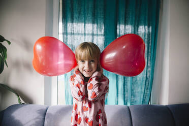 Playful girl holding balloons over ears at home - ASHF00043