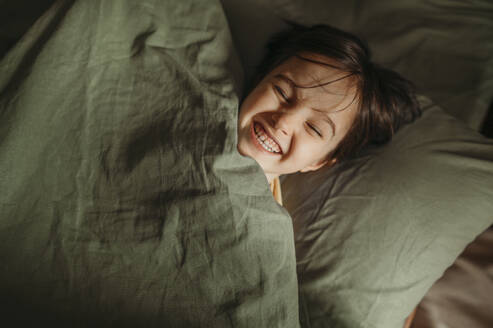 Happy boy lying under blanket in bed at home - ANAF02642