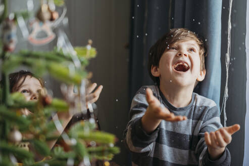 Verspielte Jungen fangen falschen Schnee in der Nähe von Weihnachtsbaum zu Hause - ANAF02652