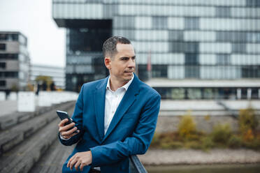 Businessman standing with smart phone in front of building - JOSEF23045