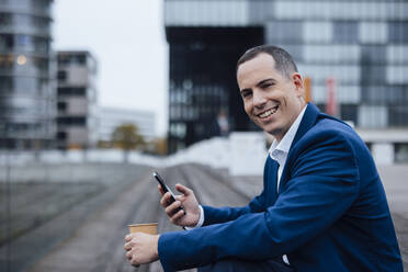 Happy businessman standing with smart phone and coffee cup in front of buildings - JOSEF23047