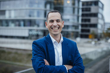 Happy businessman standing with arms crossed in front of buildings - JOSEF23053