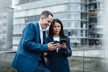 Happy businessman and businesswoman using smart phone in front of buildings - JOSEF23071