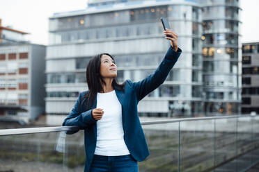 Smiling businesswoman taking selfie near railing in front of buildings - JOSEF23085