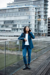 Businesswoman talking on smart phone near railing in front of buildings - JOSEF23086
