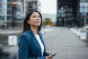 Lächelnde Geschäftsfrau, die ein Smartphone hält und im Büropark steht - JOSEF23096