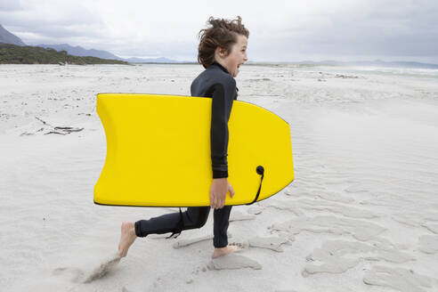 Junge (10-11) läuft am Strand und trägt ein Bodyboard - TETF02420