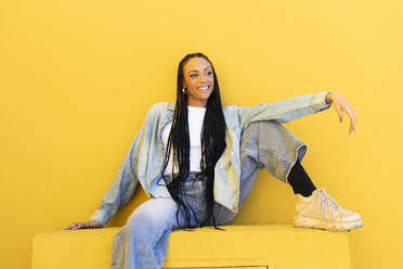 Glückliche Frau in Jeanskleidung, die in der Nähe einer gelben Wand sitzt - LMCF00763