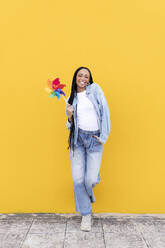 Happy woman holding pinwheel toy and leaning on yellow wall - LMCF00779