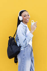 Glückliche Frau mit drahtlosen Kopfhörern vor einer gelben Wand - LMCF00783