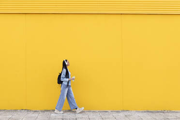 Junge Frau geht auf dem Fußweg vor einer gelben Wand - LMCF00785