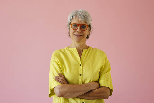 Mature woman wearing eyeglasses with arms crossed against pink background - EBSF04358