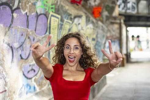 Fröhliche Frau mit Friedenszeichen in der Nähe einer Graffiti-Wand auf dem Fußweg - BFRF02441