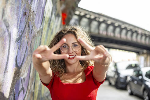 Happy woman gesturing peace sign near graffiti wall on sunny day - BFRF02442