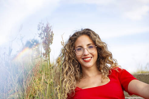Smiling young woman with curly hair wearing eyeglasses near plants - BFRF02449