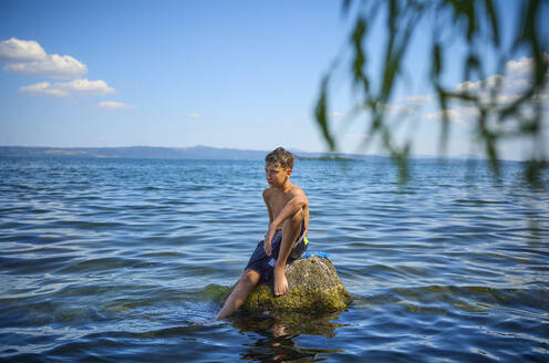 Teenage boy sitting on rock in lake Bolsena, Italy - DIKF00801