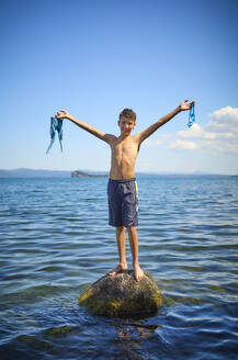 Smiling boy with arms outstretched standing on rock in lake Bolsena, Italy - DIKF00804