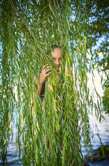 Playful girl hiding behind leaves of willow tree - DIKF00805