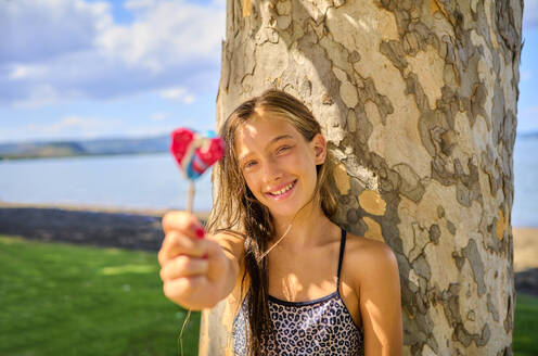 Happy girl holding lollipop near tree trunk - DIKF00813