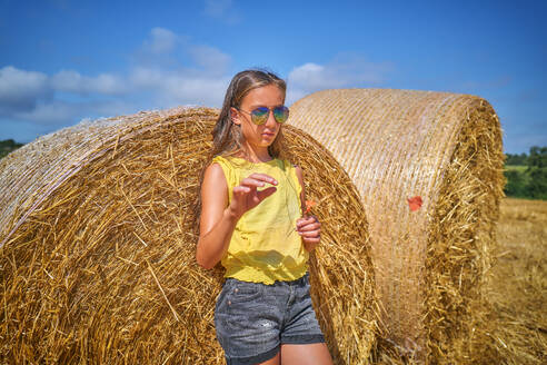 Girl wearing sunglasses and leaning on bale of straw in field - DIKF00821