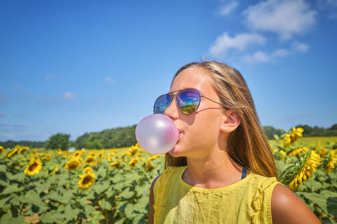 Girl blowing bubble gum in sunflower field on sunny day - DIKF00824