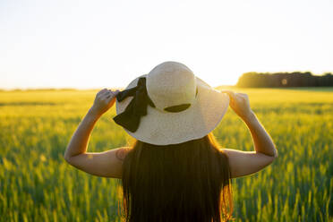 Rückansicht einer Frau mit Strohhut auf einem Feld bei Sonnenuntergang - TETF02527